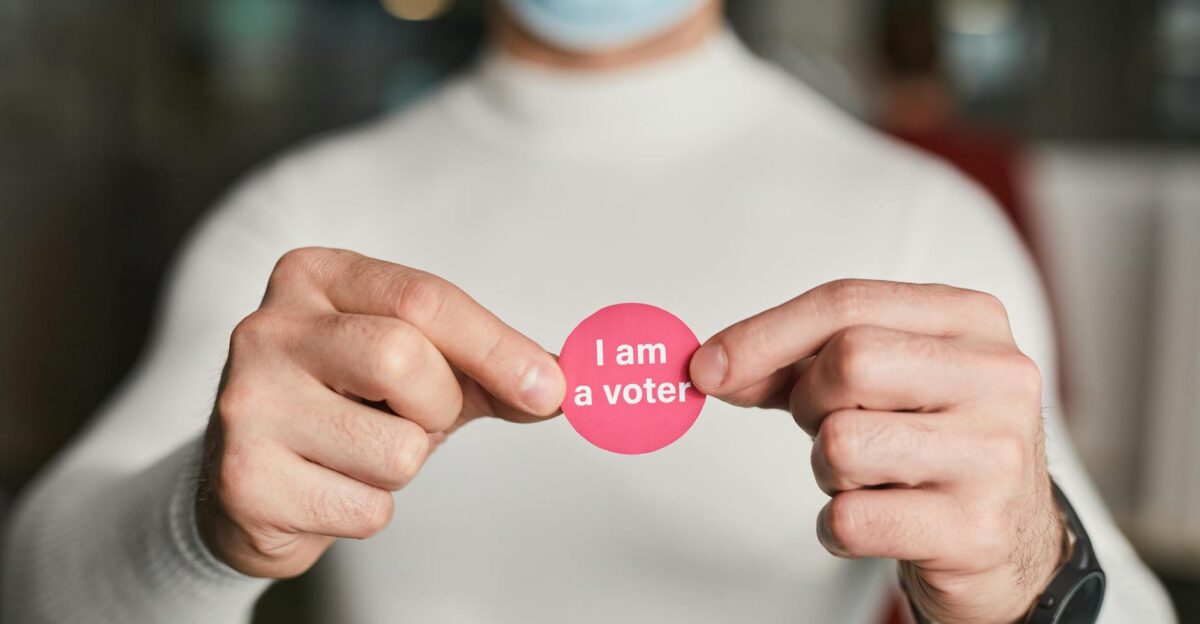 Person in a mask holding a voter sticker symbolizing civic engagement