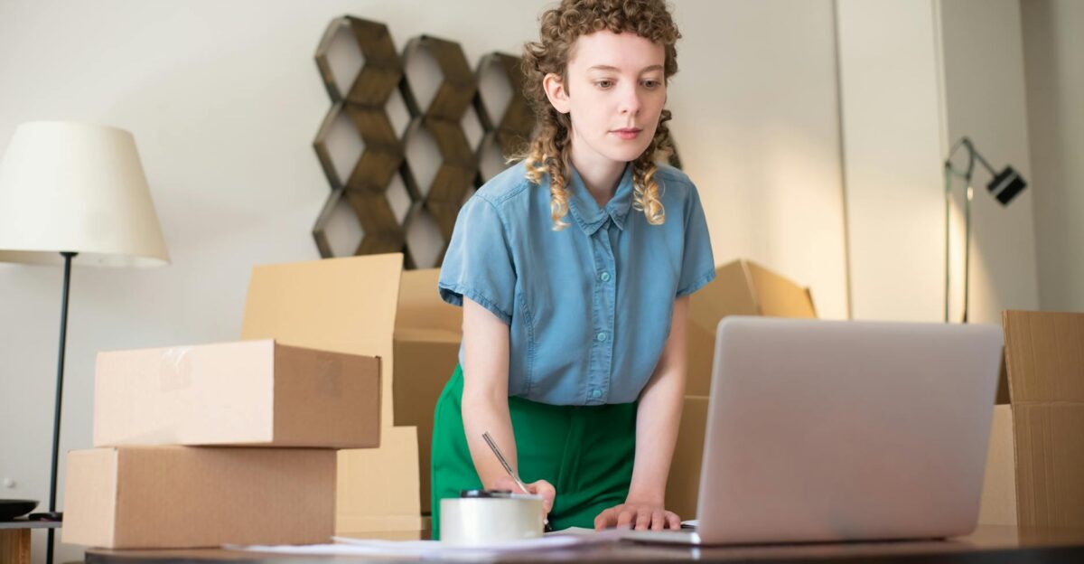 A young woman working on her laptop surrounded by cardboard boxes indicating online business operations