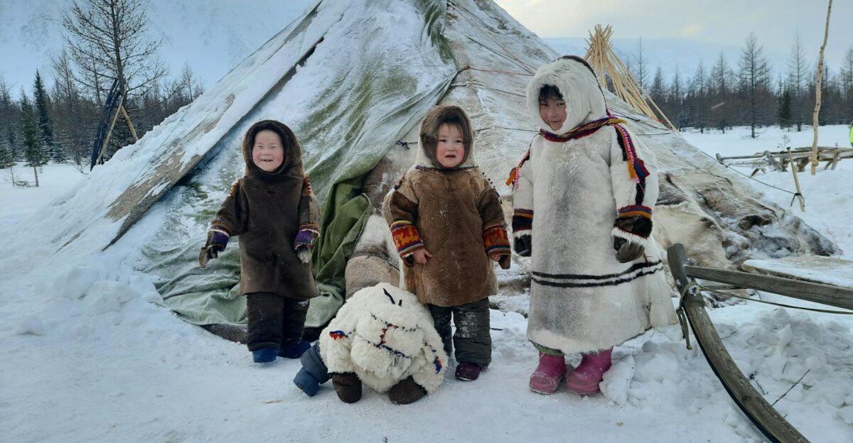 Three children in traditional Inuit clothing stand beside a snow-covered tent in a winter landscape