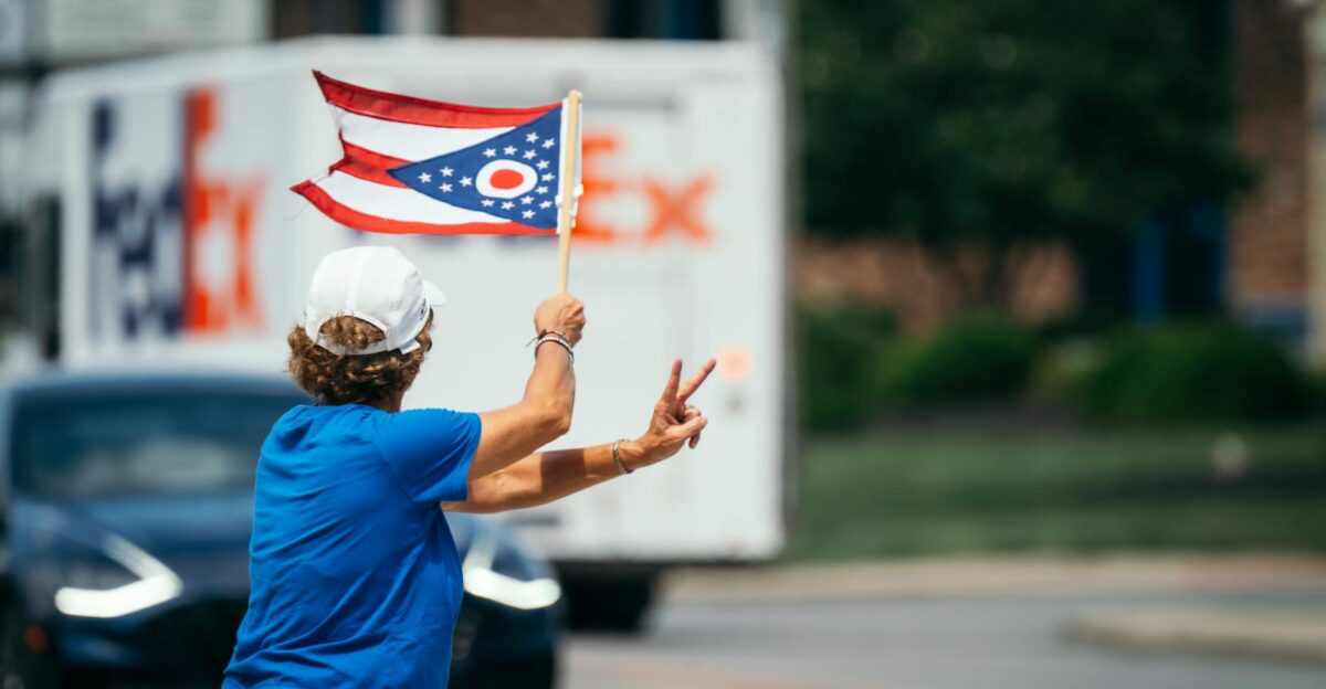 A person waves the Ohio state flag while making a peace sign on a city street